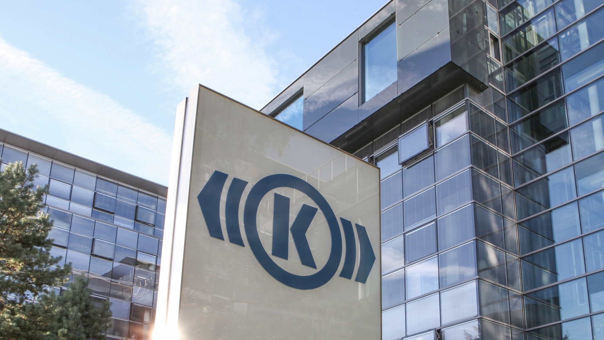 Das Foto zeigt das Knorr-Bremse Gebäude in München, in dem die Geschäftsführung der beiden Divisionen Systeme für Nutzfahrzeuge und Systeme für Schienenfahrzeuge sitzt. Im Vordergrund ein großes Eingangsschild mit dem Knorr-Bremse Logo.